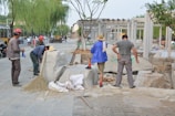 Several individuals are engaged in construction work on a paved area, surrounded by various tools and materials. Some are wearing hard hats and work attire, while others are in casual clothing. Large stone blocks and a broom are visible, along with a few vehicles in the background.