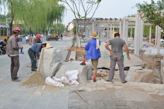 Several individuals are engaged in construction work on a paved area, surrounded by various tools and materials. Some are wearing hard hats and work attire, while others are in casual clothing. Large stone blocks and a broom are visible, along with a few vehicles in the background.