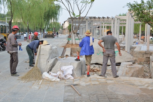 Several individuals are engaged in construction work on a paved area, surrounded by various tools and materials. Some are wearing hard hats and work attire, while others are in casual clothing. Large stone blocks and a broom are visible, along with a few vehicles in the background.