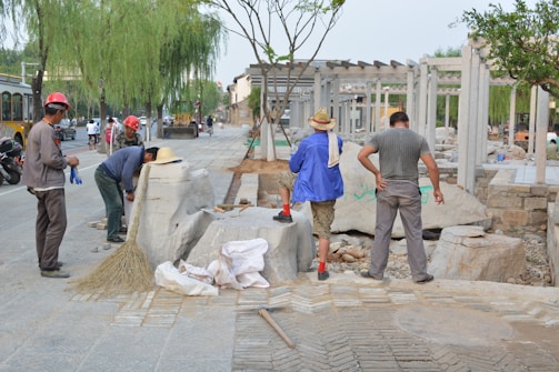Several individuals are engaged in construction work on a paved area, surrounded by various tools and materials. Some are wearing hard hats and work attire, while others are in casual clothing. Large stone blocks and a broom are visible, along with a few vehicles in the background.