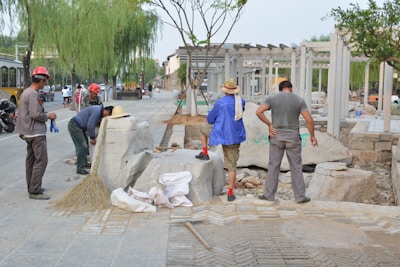 Several individuals are engaged in construction work on a paved area, surrounded by various tools and materials. Some are wearing hard hats and work attire, while others are in casual clothing. Large stone blocks and a broom are visible, along with a few vehicles in the background.