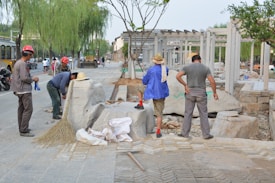 Several individuals are engaged in construction work on a paved area, surrounded by various tools and materials. Some are wearing hard hats and work attire, while others are in casual clothing. Large stone blocks and a broom are visible, along with a few vehicles in the background.