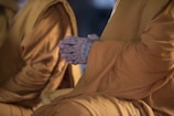 A close-up of a person wearing traditional saffron robes, with their hands clasped in a meditative position. The hands are tattooed with intricate patterns, suggesting a cultural or spiritual significance.