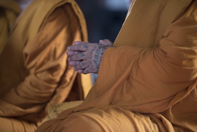 A close-up of a person wearing traditional saffron robes, with their hands clasped in a meditative position. The hands are tattooed with intricate patterns, suggesting a cultural or spiritual significance.