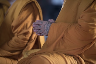 A close-up of a person wearing traditional saffron robes, with their hands clasped in a meditative position. The hands are tattooed with intricate patterns, suggesting a cultural or spiritual significance.