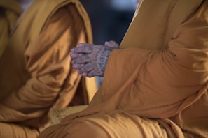 A close-up of a person wearing traditional saffron robes, with their hands clasped in a meditative position. The hands are tattooed with intricate patterns, suggesting a cultural or spiritual significance.