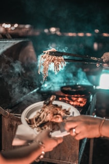 person holding black tong with grilled food in front of person holding white ceramic plate filled with grilled food