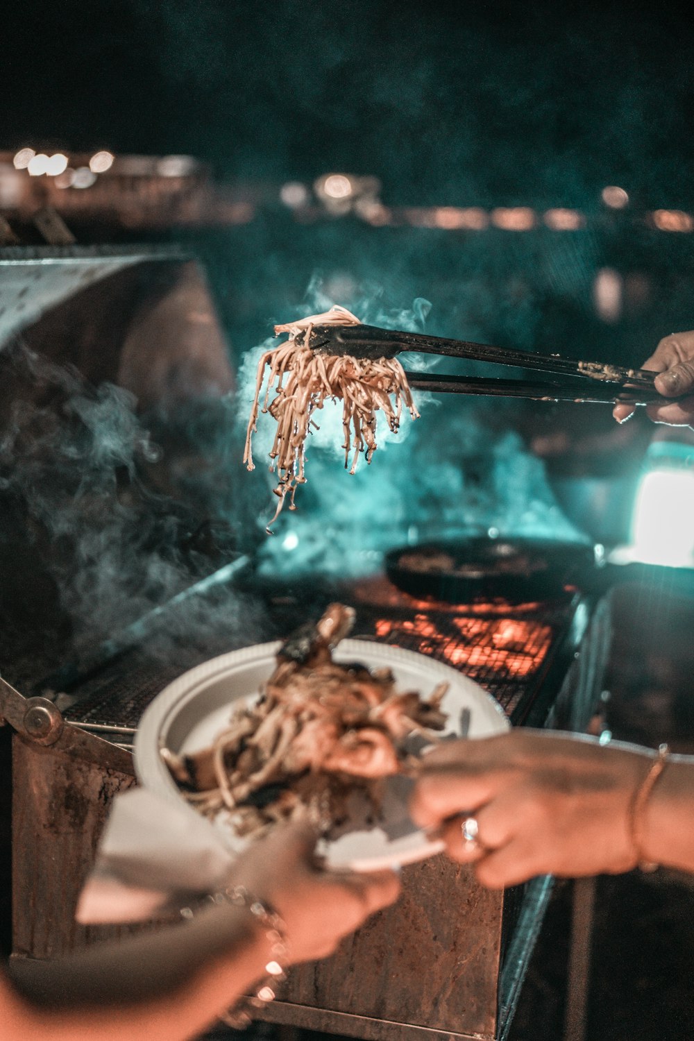 person holding black thong with grilled food in front of person holding white ceramic plate filled with grilled food