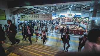 A crowded train station with numerous people walking in different directions. The scene includes staircases with yellow markings, various stalls, and signs with Japanese text. People are dressed in business attire, indicating a busy commute or market setting.