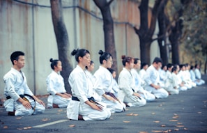 A group of people dressed in white martial arts uniforms are kneeling in meditation or focus. They are positioned in a row on an outdoor pathway surrounded by trees and a concrete wall.