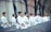 A group of people dressed in white martial arts uniforms are kneeling in meditation or focus. They are positioned in a row on an outdoor pathway surrounded by trees and a concrete wall.