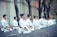 A group of people dressed in white martial arts uniforms are kneeling in meditation or focus. They are positioned in a row on an outdoor pathway surrounded by trees and a concrete wall.
