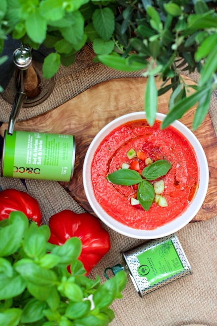 A bowl of vibrant red gazpacho garnished with fresh basil leaves and diced cucumbers is placed on a wooden surface. Surrounding the bowl are fresh red bell peppers, a bright green canister, and lush green herbs, creating a fresh and natural setting.
