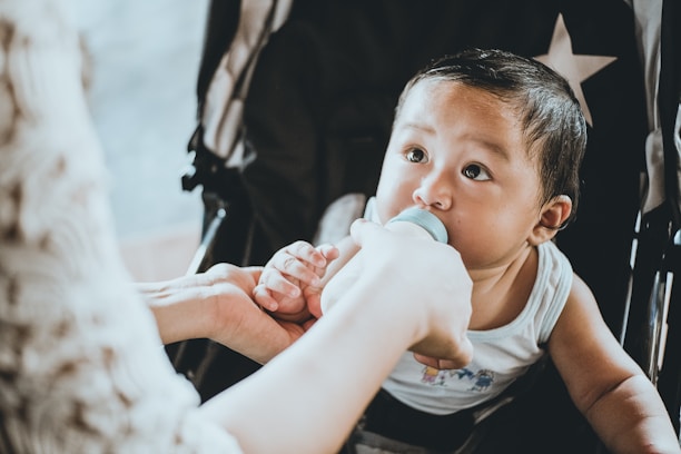 A baby is being bottle-fed in a stroller. The child is wearing a sleeveless top and appears to be gazing up while holding onto the bottle. The scene is intimate and nurturing, suggesting a moment of care and attention. There are gentle shadows, and the star pattern on the stroller's fabric is visible.