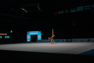 A gymnast performs a floor routine with a ribbon, showcasing a handstand in a dimly lit indoor arena. The focus is on the gymnast's skill and the vibrant colors of the ribbon they are maneuvering.