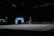 A gymnast performs a floor routine with a ribbon, showcasing a handstand in a dimly lit indoor arena. The focus is on the gymnast's skill and the vibrant colors of the ribbon they are maneuvering.