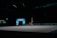 A gymnast performs a floor routine with a ribbon, showcasing a handstand in a dimly lit indoor arena. The focus is on the gymnast's skill and the vibrant colors of the ribbon they are maneuvering.