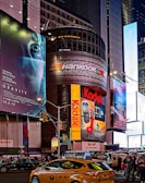 A bustling urban scene features towering buildings adorned with large, colorful advertisements and billboards. Brands like Kodak and Hankook are prominently displayed. The scene is set in a busy street intersection with numerous pedestrians and yellow taxis navigating through the area, creating a vibrant city atmosphere.