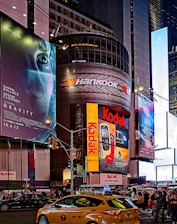 A bustling urban scene features towering buildings adorned with large, colorful advertisements and billboards. Brands like Kodak and Hankook are prominently displayed. The scene is set in a busy street intersection with numerous pedestrians and yellow taxis navigating through the area, creating a vibrant city atmosphere.