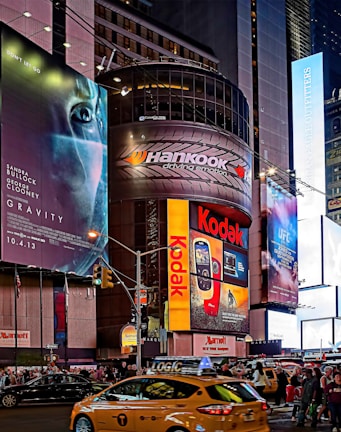 A bustling urban scene features towering buildings adorned with large, colorful advertisements and billboards. Brands like Kodak and Hankook are prominently displayed. The scene is set in a busy street intersection with numerous pedestrians and yellow taxis navigating through the area, creating a vibrant city atmosphere.