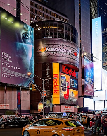 A bustling urban scene features towering buildings adorned with large, colorful advertisements and billboards. Brands like Kodak and Hankook are prominently displayed. The scene is set in a busy street intersection with numerous pedestrians and yellow taxis navigating through the area, creating a vibrant city atmosphere.