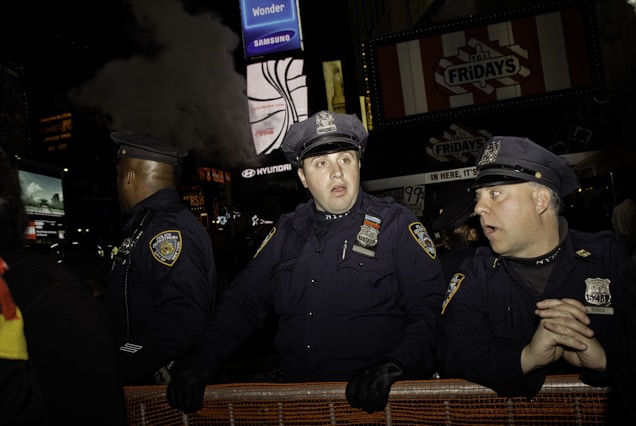 A group of police officers stands in front of a barricade, appearing alert and focused. They are wearing dark uniforms with badges and hats, illuminated by the ambient light of the city at night. In the background, bright signs from businesses like TGI Fridays and advertisements create a lively atmosphere typical of a bustling city environment.