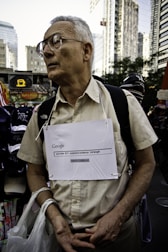 An older man wearing glasses stands in an urban environment, possibly a city street surrounded by tall buildings. He is wearing a beige shirt and has a handmade sign around his neck with 'Google' and an internet search query related to '9/11 explosive evidence' written on it. He carries plastic bags in one hand and appears contemplative or serious.