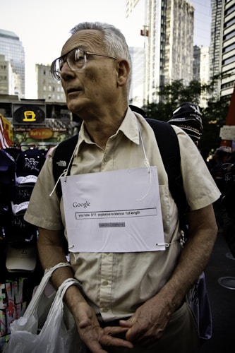 An older man wearing glasses stands in an urban environment, possibly a city street surrounded by tall buildings. He is wearing a beige shirt and has a handmade sign around his neck with 'Google' and an internet search query related to '9/11 explosive evidence' written on it. He carries plastic bags in one hand and appears contemplative or serious.