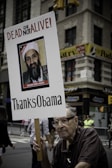 An elderly man holding a protest sign with an image of Osama Bin Laden that reads 'DEAD OR ALIVE' and 'Thanks Obama' in a city street setting. There are buildings in the background and a few people visible behind barricades.