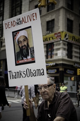 An elderly man holding a protest sign with an image of Osama Bin Laden that reads 'DEAD OR ALIVE' and 'Thanks Obama' in a city street setting. There are buildings in the background and a few people visible behind barricades.