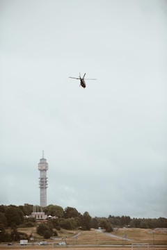 A helicopter is flying in a cloudy sky over a landscape with a distinctive tall tower surrounded by trees and grassy fields. Below, a road and various buildings are visible.