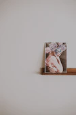A framed photograph is placed on a simple wooden shelf. The image within the frame shows a person with intricate floral headpieces and face paint, featuring soft pastel colors. The background is a plain, light-colored wall that enhances the minimalistic and artistic presentation.