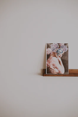 A framed photograph is placed on a simple wooden shelf. The image within the frame shows a person with intricate floral headpieces and face paint, featuring soft pastel colors. The background is a plain, light-colored wall that enhances the minimalistic and artistic presentation.