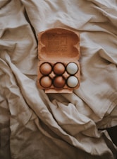 A cardboard carton contains six eggs with varying shades of brown and one white egg, resting on a crumpled, textured fabric with soft lighting.