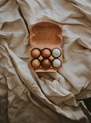 A cardboard carton contains six eggs with varying shades of brown and one white egg, resting on a crumpled, textured fabric with soft lighting.