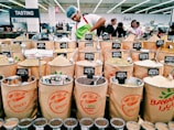 A market scene with various bags filled with different types of spices displayed for sale. Each bag has a label showing the type of spice and its price. In the background, a person wearing a white shirt and a hairnet is working behind the spice counter. Several customers are visible in the background, amidst bright lighting and multilingual signs indicating different sections.