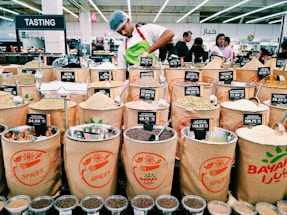 A market scene with various bags filled with different types of spices displayed for sale. Each bag has a label showing the type of spice and its price. In the background, a person wearing a white shirt and a hairnet is working behind the spice counter. Several customers are visible in the background, amidst bright lighting and multilingual signs indicating different sections.