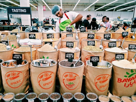 A market scene with various bags filled with different types of spices displayed for sale. Each bag has a label showing the type of spice and its price. In the background, a person wearing a white shirt and a hairnet is working behind the spice counter. Several customers are visible in the background, amidst bright lighting and multilingual signs indicating different sections.