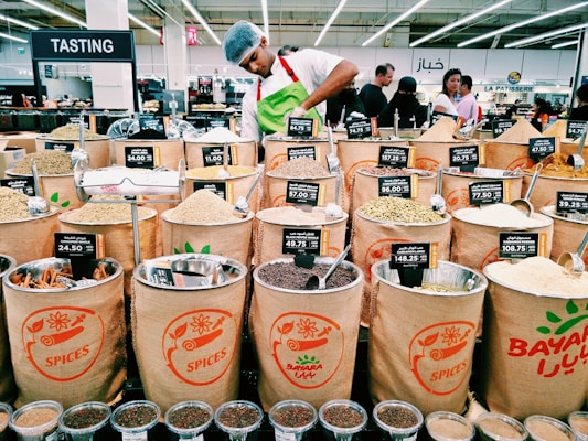 A market scene with various bags filled with different types of spices displayed for sale. Each bag has a label showing the type of spice and its price. In the background, a person wearing a white shirt and a hairnet is working behind the spice counter. Several customers are visible in the background, amidst bright lighting and multilingual signs indicating different sections.