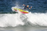 A surfer skillfully rides a wave, performing a maneuver on a vibrant, yellow and red surfboard. The ocean water splashes around as the surfer maintains balance with one arm raised in a peace sign.