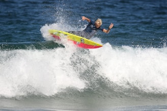 A surfer skillfully rides a wave, performing a maneuver on a vibrant, yellow and red surfboard. The ocean water splashes around as the surfer maintains balance with one arm raised in a peace sign.