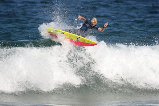 A surfer skillfully rides a wave, performing a maneuver on a vibrant, yellow and red surfboard. The ocean water splashes around as the surfer maintains balance with one arm raised in a peace sign.