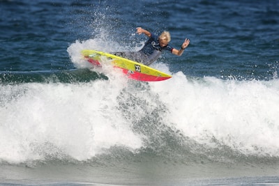 A surfer skillfully rides a wave, performing a maneuver on a vibrant, yellow and red surfboard. The ocean water splashes around as the surfer maintains balance with one arm raised in a peace sign.