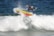 A surfer skillfully rides a wave, performing a maneuver on a vibrant, yellow and red surfboard. The ocean water splashes around as the surfer maintains balance with one arm raised in a peace sign.
