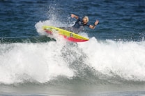 A surfer skillfully rides a wave, performing a maneuver on a vibrant, yellow and red surfboard. The ocean water splashes around as the surfer maintains balance with one arm raised in a peace sign.