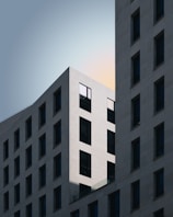 A modern architectural design featuring two intersecting buildings with a minimalist style. The buildings have a grid-like pattern of windows, and the lighting emphasizes sharp lines and geometric shapes. The sky in the background is soft with subtle hues, highlighting the clean and elegant structure.