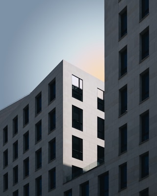 A modern architectural design featuring two intersecting buildings with a minimalist style. The buildings have a grid-like pattern of windows, and the lighting emphasizes sharp lines and geometric shapes. The sky in the background is soft with subtle hues, highlighting the clean and elegant structure.