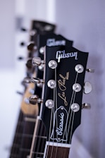 A close-up view of the headstocks of several guitars, focusing on a 'Gibson Les Paul Classic' model with chrome tuning pegs and a glossy finish. The guitars are aligned in a row against a neutral background, highlighting the craftsmanship and branding.