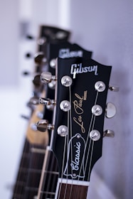 A close-up view of the headstocks of several guitars, focusing on a 'Gibson Les Paul Classic' model with chrome tuning pegs and a glossy finish. The guitars are aligned in a row against a neutral background, highlighting the craftsmanship and branding.