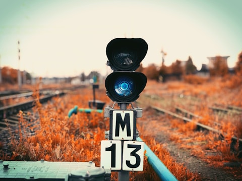 A railway signal light stands prominently in the foreground with a vivid blue light illuminated. The surrounding area features railway tracks trailing off into the blurred background and autumnal vegetation with a warm, earthy hue.